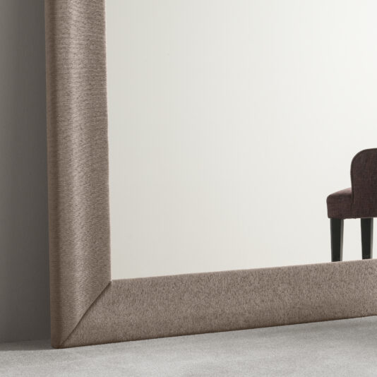 The Large Contemporary Fabric Floor Mirror, featuring a textured beige frame, is positioned against a light-colored wall. A section of a dark upholstered chair can be seen in the reflection on the right side of the mirror. The floor is covered with light gray carpet.