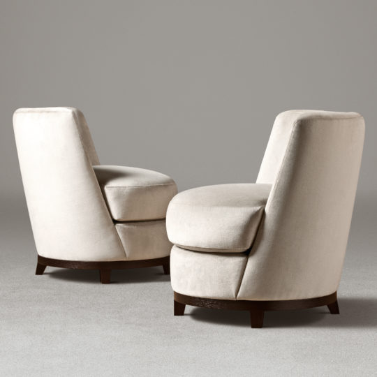 Two Curved Occasional Chairs With Swivel Bases, featuring modern beige upholstery and a round design, are positioned facing each other against a plain grey background. These chairs offer comfortable and stylish cushioned seating.