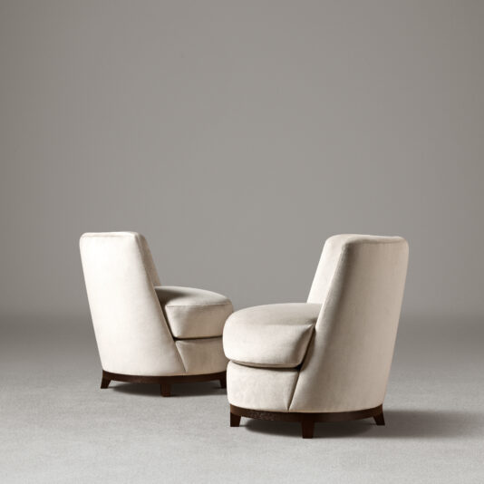 Curved Occasional Chair With Swivel Base Two Curved Occasional Chairs With Swivel Bases featuring beige, cushioned seats sit angled toward each other against a neutral gray background. The soft, plush texture of the upholstery adds a touch of comfort. These barrel chairs with wooden bases create a minimalist setting, highlighting their sleek and modern design.