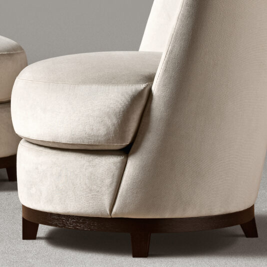 Curved Occasional Chair With Swivel Base A close-up of the Curved Occasional Chair With Swivel Base showcases its beige upholstery, rounded design, and cushioned backrest. The chair features a sturdy dark wooden base with short legs, adding to its modern and elegant appearance. Its swivel base enhances versatility, set against a background blurred in gray tones.