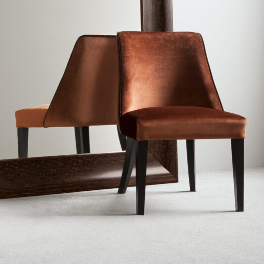 A Velvet High End Dining Chair with dark wooden legs is positioned in front of a large mirror. The chair's reflection is visible in the mirror, creating the illusion of two chairs. The setting has neutral-colored walls and a plush, light-colored carpet.