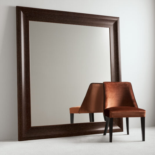 A large, dark wooden framed mirror leaning against a light gray wall reflects the Velvet High End Dining Chair with black legs. The reflection shows the chair from the back, highlighting the texture and sheen of its velvet fabric. The room has a clean, minimalist ambiance.