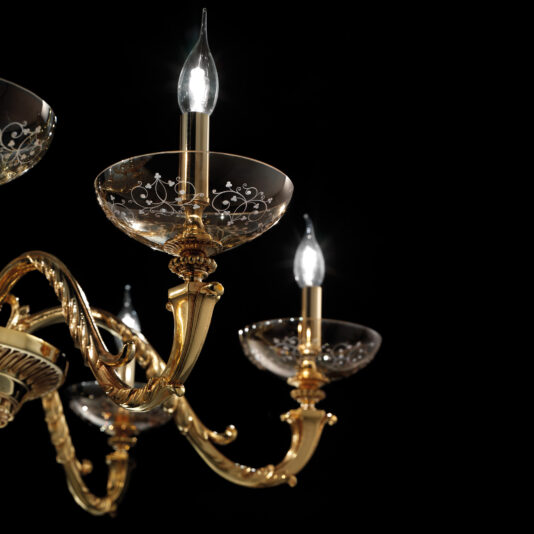 Amber Hand Blown Glass Chandelier A close-up of the Amber Hand Blown Glass Chandelier reveals its ornate gold structure adorned with three lit candle-shaped bulbs. The chandelier features intricate details and delicate hand-blown glass bowls, each etched with floral patterns, all set against a dark background.