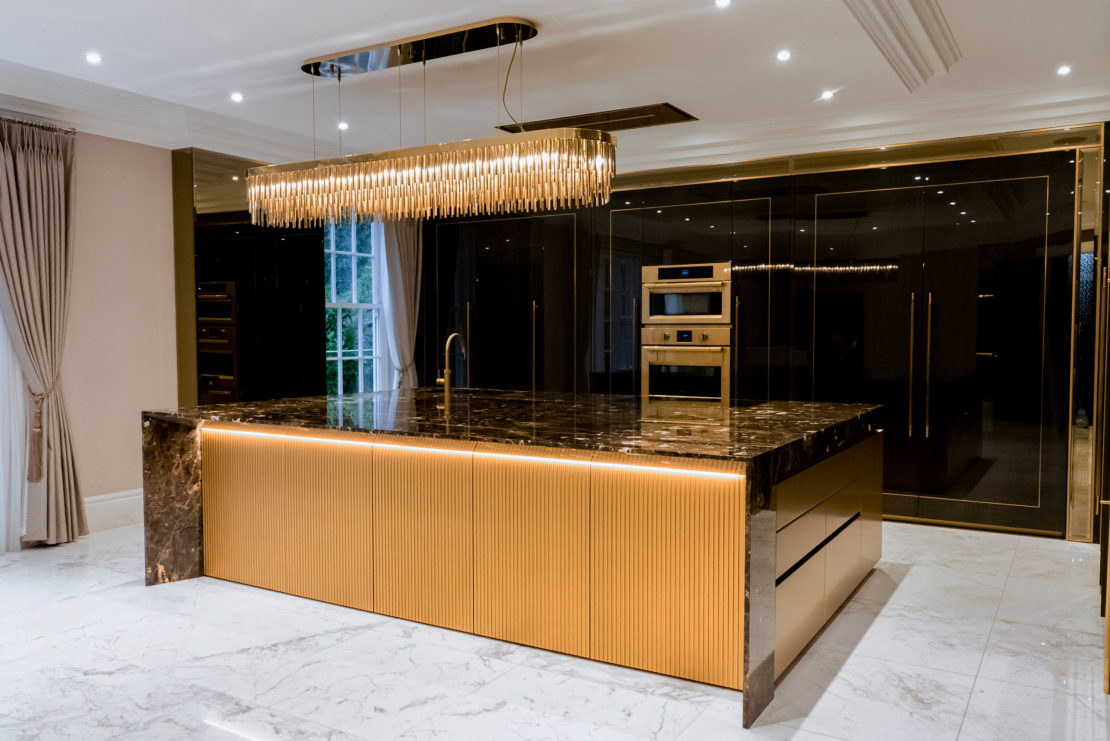 A modern luxury family kitchen featuring a large marble-topped island with a gold base. Above the island is an elegant chandelier with hanging crystals. The kitchen includes built-in stainless steel appliances, dark glossy cabinets, and white marble flooring.