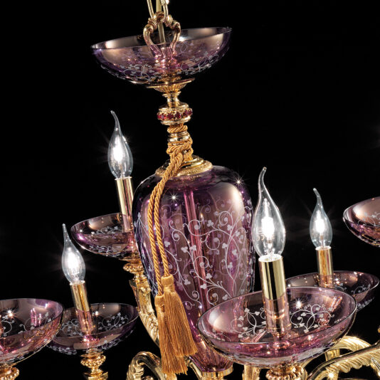 The Hand Blown Amethyst Glass Chandelier is a luxurious lighting fixture adorned with intricate golden details and purple crystal accents. The chandelier features multiple bulbs designed to resemble candle flames, delicate floral patterns on the crystals, and golden tassels hanging from the center structure.