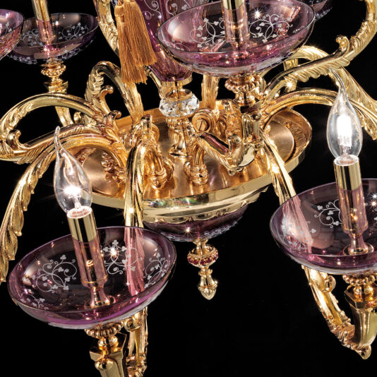 The Hand Blown Amethyst Glass Chandelier boasts a golden, ornate design with multiple arms that hold transparent amethyst glass cups. Each hand-blown cup is enhanced with elegant white patterns and some cradle glowing candle-shaped light bulbs, creating a striking contrast against a black background.