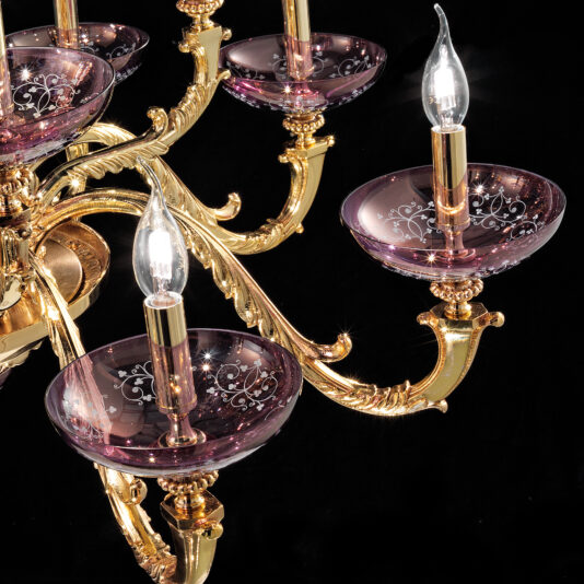 The Hand Blown Amethyst Glass Chandelier features golden metallic arms and delicate hand-blown amethyst glass candle holders, each containing a lit bulb designed to resemble a candle flame. The purple glass showcases intricate white floral designs while the chandelier is displayed against a black background.