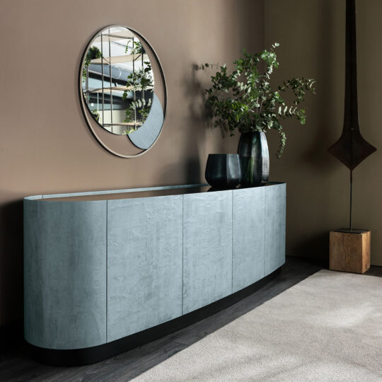 A sleek, modern Blue Slate Effect Four Door Buffet with a curved design and a textured light gray finish stands against a beige wall. Atop the buffet are two tall black vases filled with green foliage, while above it hangs a round mirror reflecting the opposite room.