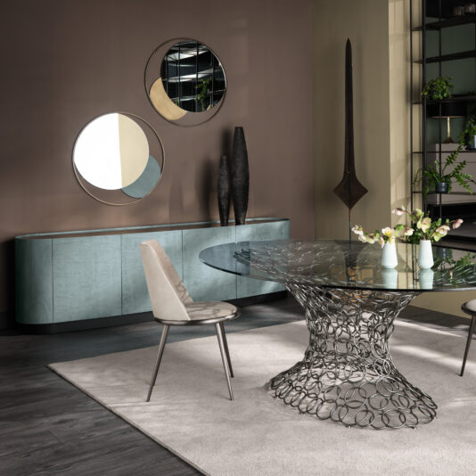 Blue Slate Effect Four Door Buffet A chic dining room showcasing a glass table with a sculptural metal base, complemented by a cushioned chair and a light gray rug. The back wall is adorned with the Blue Slate Effect Four Door Buffet, three round mirrors, and two tall decorative vases. In the background, shelves display an assortment of potted plants.