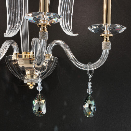 Triple Leaf Wall Light With Swarovski® Crystals A close-up of an elegant chandelier with crystal and glass components. The design includes curving transparent arms, adorned with faceted crystal drops hanging from each arm. The background, reminiscent of the Triple Leaf Wall Light With Swarovski® Crystals, features a dark, plain surface that highlights the chandelier's intricate details.