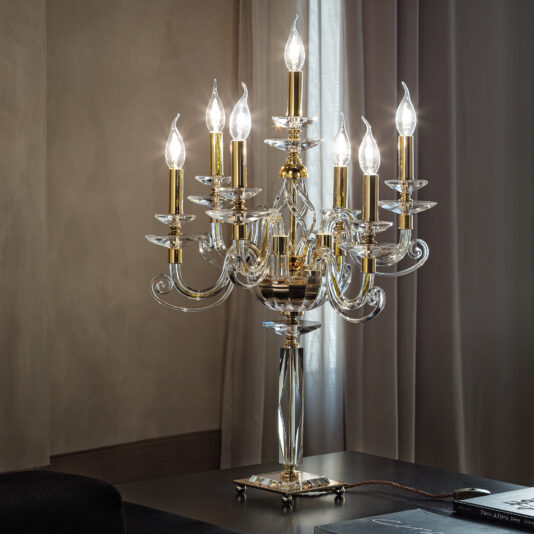 The Candle Chandelier Style Table Lamp, adorned with decorative crystal and brass, casts a warm glow on a dark wooden table in a dimly lit room. The eight-arm lamp, featuring candle-like light bulbs, is beautifully set against heavy grey curtains.