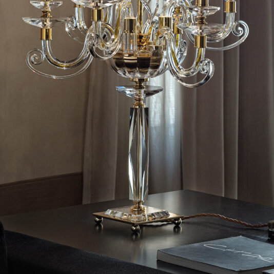 The Candle Chandelier Style Table Lamp, featuring gold accents and multiple glass arms, rests elegantly on a dark table. Its intricately designed base with crystal elements highlights its luxurious craftsmanship against a backdrop of grey curtains and an open book.