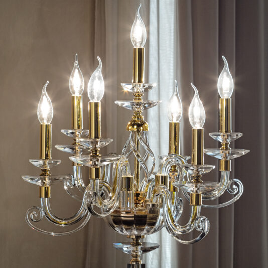 A close-up of the Candle Chandelier Style Table Lamp showcases its seven lit candle-shaped bulbs with clear glass and gold accents. The lamp's elegant design includes spiral details, echoing the sophisticated style of a chandelier, set against a neutral background with draped curtains.