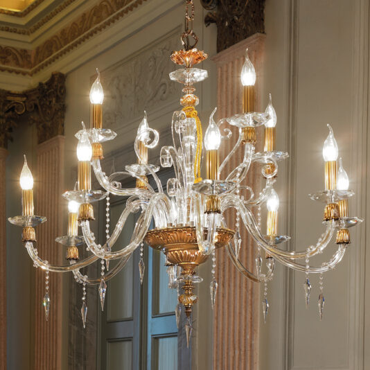 A Candle Style Chandelier adorned with multiple lit bulbs and intricate glasswork hangs from the ceiling. This chandelier showcases gold and crystal elements, featuring decorative curving arms and teardrop-shaped pendants. The background reveals part of an elegant room with ornate moldings.
