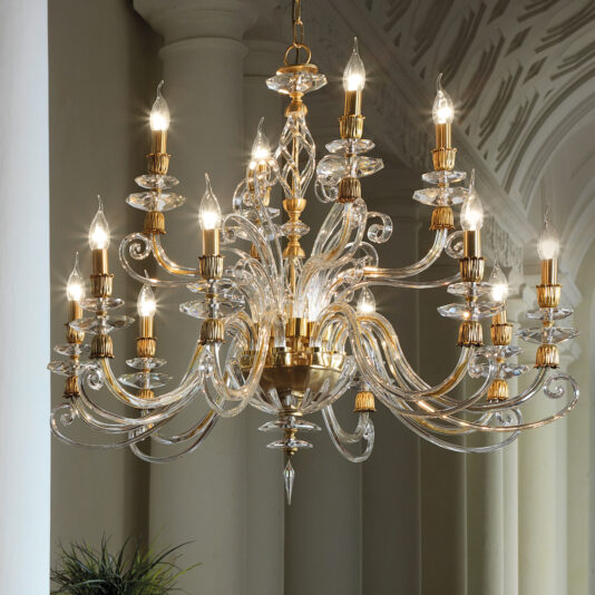 The **Candle Style Swarovski® Crystal Chandelier** is a grand, ornate piece featuring multiple tiers of curved, clear glass arms holding candle-like light bulbs with golden accents. The chandelier stands out beautifully against a backdrop of white, columned arches and an intricately patterned ceiling.