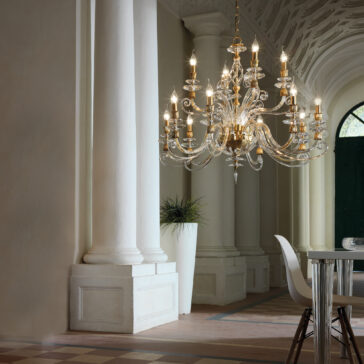 Candle Style Swarovski® Crystal Chandelier A grand, ornate Candle Style Swarovski® Crystal Chandelier with lit candles hangs in an elegant room featuring white columns and intricate ceiling details. The space is adorned with a large potted plant and modern furniture, including a white chair and table.