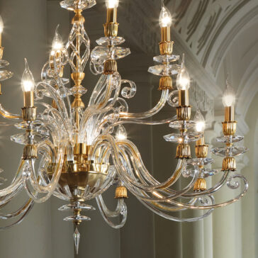 Candle Style Swarovski® Crystal Chandelier A close-up view of the Candle Style Swarovski® Crystal Chandelier, featuring an ornate design with multiple curved arms and candle-style light bulbs. Adorned with stunning Swarovski® crystals, this gold chandelier casts a soft, warm glow. The elegant fixture hangs from a ceiling with intricate molding details in a sophisticated interior setting.