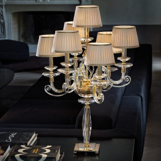 The Classic Chandelier Style Table Lamp, featuring multi-armed crystal accents and pleated cream-colored lampshades, elegantly graces a dark table. The sophisticated setting is enhanced by dark furniture, a decorative fireplace, and neatly placed books on the table.