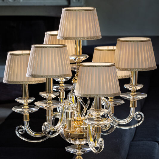 A detailed view of the Classic Chandelier Style Table Lamp reveals its exquisite, multi-arm crystal design adorned with beige pleated lampshades. This sophisticated table lamp showcases intricately curved glass arms and luxurious gold accents, all beautifully illuminated against a dark, blurred background.