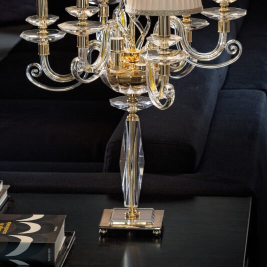 A luxurious Classic Chandelier Style Table Lamp, featuring intricate crystal and gold details, sits on a dark coffee table. In the background is a black sofa adorned with plush cushions. Beside the lamp on the table lies a hardcover book with an abstract cover design.
