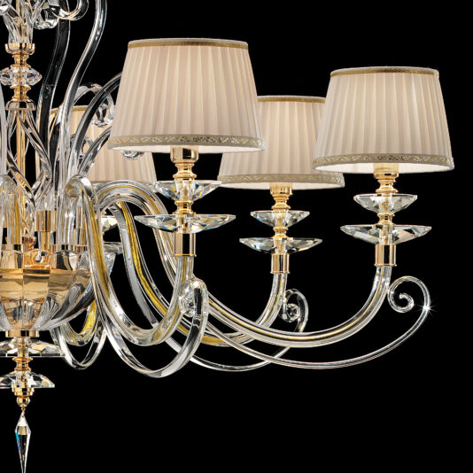 Classic Style Chandelier With Swarovski® Crystals A close-up of the Classic Style Chandelier with Swarovski® Crystals showcases its elegant, classic design, featuring multiple crystal arms and light fixtures. Each fixture is adorned with a pleated beige lampshade with gold trim. The chandelier's detailed crystal and gold accents glimmer beautifully against a black background.
