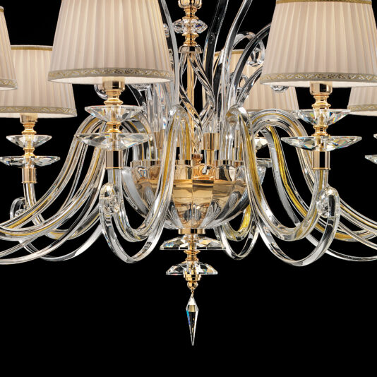 Classic Style Chandelier With Swarovski® Crystals A close-up of the Classic Style Chandelier With Swarovski® Crystals reveals its ornate, classic design with multiple curved glass arms radiating from the center. Each arm supports a decorative gold stem crowned with a lampshade crafted from pleated cream fabric and embellished with Swarovski® crystals. The intricate chandelier stands out against a black background.