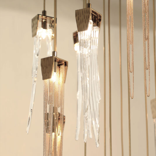 The Contemporary Chain And Glass Stairwell Chandelier features modern vertical glass elements and gold chains hanging from the ceiling. It emits a soft, warm glow that creates an elegant and sophisticated ambiance, capturing a minimalist aesthetic.
