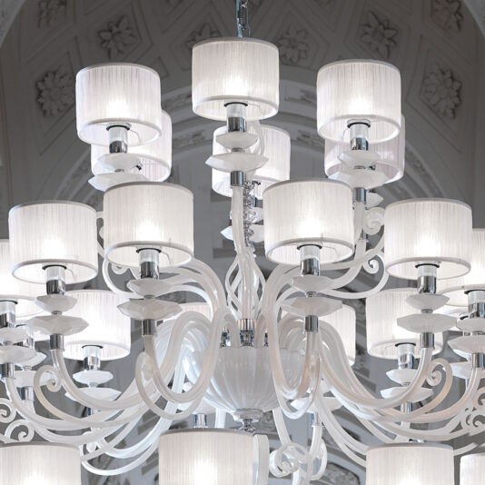 A close-up of the Large White Glass Modern Chandelier reveals its intricate arrangement of multiple white glass lampshades. This ornate chandelier showcases a swirling arm design in white and hangs against an intricately patterned, light-colored ceiling.