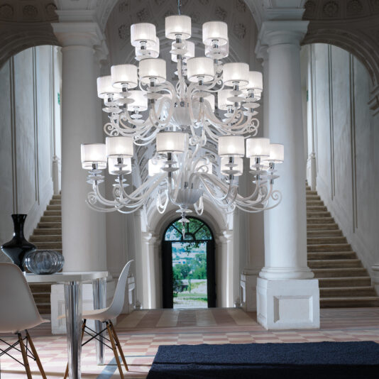 A Large White Glass Modern Chandelier with intricate details hangs majestically in a grand hall adorned with high ceilings, white columns, and two symmetrical staircases. Below it rests a contemporary seating area highlighted by a round glass table surrounded by white chairs and complemented by decorative vases.