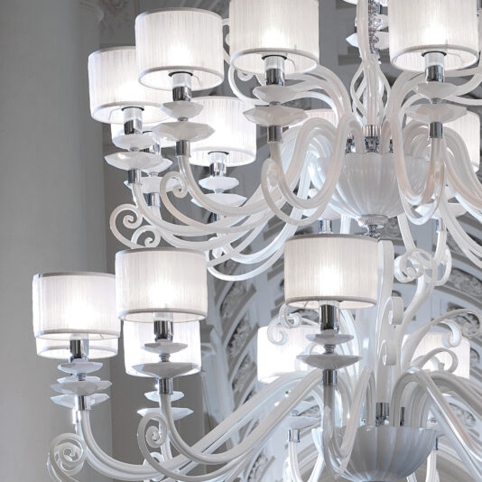 The Large White Glass Modern Chandelier, with its luxurious and expansive design, features multiple tiers adorned with white fabric lampshades. Its elaborate, swirling arms add an elegant touch as it hangs gracefully against an intricately decorated white background in a sophisticated interior space.