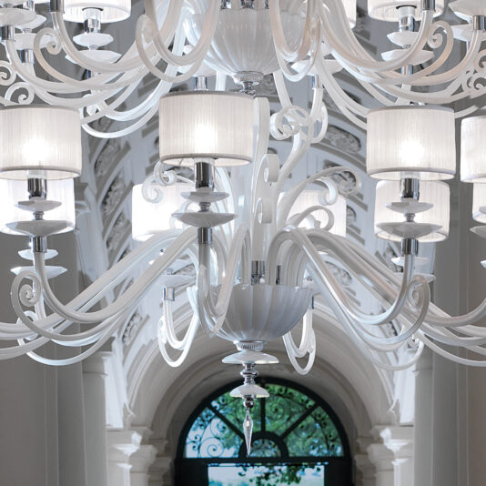 The Large White Glass Modern Chandelier, adorned with multiple lampshades, intricate curving arms, and hanging decorative pieces, sets a sophisticated tone from its high, arched ceiling with ornate accents in a well-lit room. Natural light filters through a large arched window in the background.