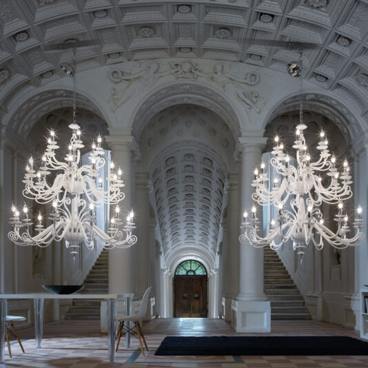 A grand hall with an ornate, vaulted ceiling, carved stone details, and a symmetrical staircase leading to an arched doorway. Two Large White Glass Modern Chandeliers hang from the ceiling. A contemporary white table and chairs are placed to the left.