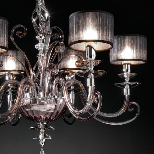 The Modern Smoked Glass Chandelier boasts a luxurious design featuring intricate glass arm motifs and six dark, translucent lampshades. These lampshades allow light to diffuse gently, creating a soft glow. Set against a dark backdrop, the chandelier's elegant appearance is further enhanced.