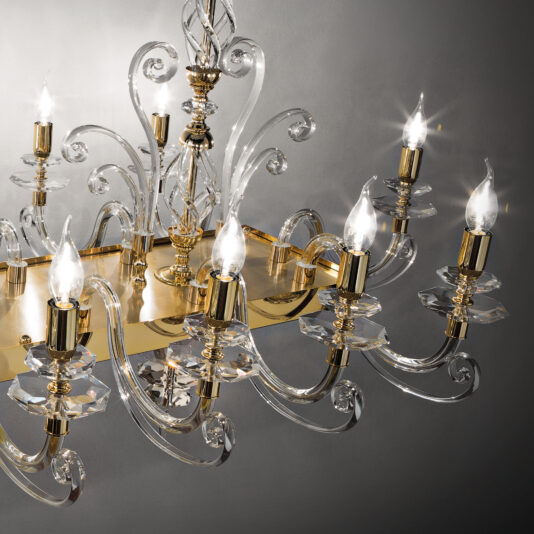 A close-up of the Rectangular Classic Candle Style Chandelier showcases its multiple curving glass arms, each elegantly holding a lit candle-shaped bulb. The chandelier is adorned with gold accents and intricate detailing, set against a plain gray background.