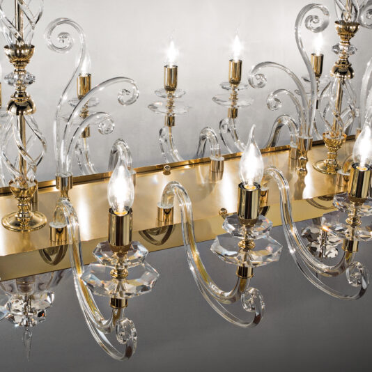 The Rectangular Classic Candle Style Chandelier boasts multiple candle-shaped bulbs and ornate gold accents. It features intricate transparent glass arms and decorative curves that elegantly reflect light, all displayed against a neutral background.