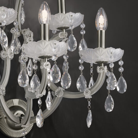 Close-up of the Satin Glass Classic Style Wall Light with lit candle-shaped bulbs. The fixture features white, decorative floral designs and numerous hanging crystal prisms that reflect the light. Satin glass elements add an extra touch of elegance. The dark background creates a striking contrast with the illuminated wall light.