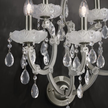 Close-up of a Satin Glass Classic Style Wall Light with crystal pendants and white, ornate floral details against a dark background. The fixture features satin glass elements and multiple curved arms, each adorned with hanging crystals and lit bulbs at the top, emitting a soft, warm light.