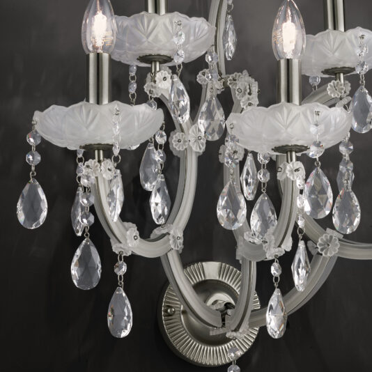 Close-up of a Satin Glass Classic Style Wall Light with crystal pendants and white, ornate floral details against a dark background. The fixture features satin glass elements and multiple curved arms, each adorned with hanging crystals and lit bulbs at the top, emitting a soft, warm light.