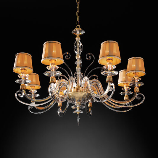 The Swarovski® Crystal Chandelier With Shades exudes luxurious, classic style with its intricate, curving glass arms and gold accents. Featuring eight amber-colored lampshades adorned with Swarovski® crystals, this chandelier is suspended by a gold chain against a dark background, illuminating the space with a warm, inviting glow.