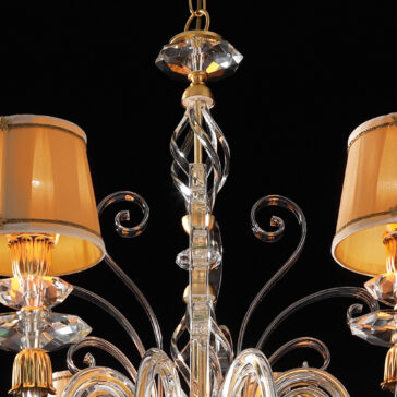 Swarovski® Crystal Chandelier With Shades A close-up view of the Swarovski® Crystal Chandelier With Shades reveals its classic style, highlighted by Swarovski® crystals and gold detailing. The fixture boasts multiple arms with elegant scrollwork, adorned with candle-style lights, each covered by a light brown shade. The black background further accentuates the chandelier's intricate design.