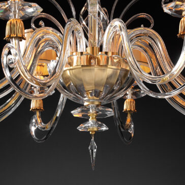 Swarovski® Crystal Chandelier With Shades A close-up shot of the Swarovski® Crystal Chandelier With Shades reveals its elegant, classic style with gold accents and intricate, curved glass arms. The chandelier features multiple lights, each adorned with individual gold shades and sparkling Swarovski® crystals, creating a luxurious and sophisticated appearance against a dark background.