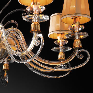 Swarovski® Crystal Chandelier With Shades A close-up view of the Swarovski® Crystal Chandelier With Shades showcases multiple lampshades. The chandelier flaunts intricately curved, clear glass arms adorned with Swarovski® crystals. The warm light emanating from the lampshades creates a luxurious and sophisticated ambiance against a dark background.