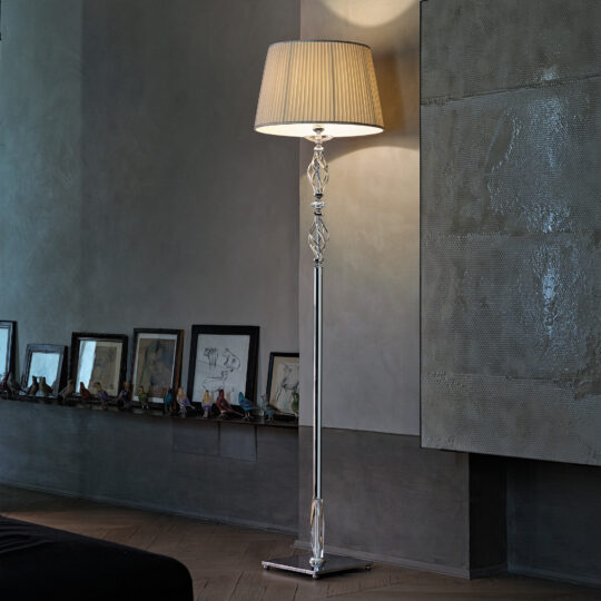 A sleek, contemporary room featuring the Twisted Floor Lamp With Swarovski® Crystal and a beige lampshade casting a soft light. The wall behind showcases framed artwork and decorative items displayed on a long shelf, with textured artwork hanging above.