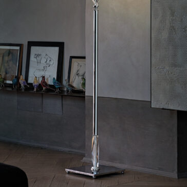 Twisted Floor Lamp With Swarovski® Crystal A sleek, modern Twisted Floor Lamp With Swarovski® Crystal stands in a dimly lit room. Behind it, there’s a shelf holding framed artwork and small, colorful bird figurines. The wooden floor complements the textured, reflective surface of the wall to the right. This twisted floor lamp features Swarovski® Crystal accents that catch the light beautifully.