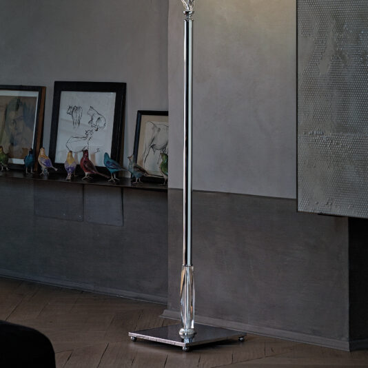 Twisted Floor Lamp With Swarovski® Crystal A sleek, modern Twisted Floor Lamp With Swarovski® Crystal stands in a dimly lit room. Behind it, there’s a shelf holding framed artwork and small, colorful bird figurines. The wooden floor complements the textured, reflective surface of the wall to the right. This twisted floor lamp features Swarovski® Crystal accents that catch the light beautifully.