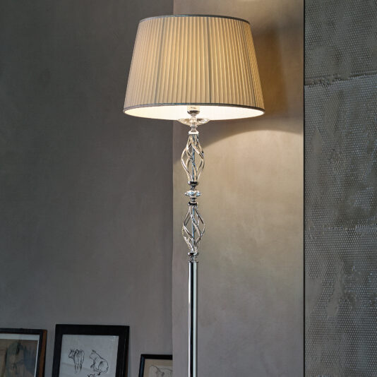 Twisted Floor Lamp With Swarovski® Crystal The Twisted Floor Lamp With Swarovski® Crystal, boasting an intricate metallic stand and a pleated beige lampshade, is set beside a wall decorated with framed artwork. The lamp emits a warm, soft light, enhancing the room's cozy ambiance.