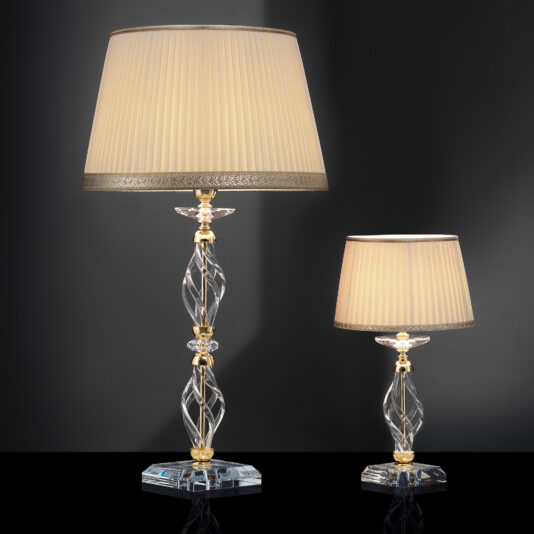 An image showcases two Twisted Table Lamps With Swarovski® Crystal, each featuring beige pleated shades and intricately designed glass bases adorned with Swarovski® Crystal accents. The taller lamp is positioned on the left, while the shorter one sits on the right. Both lamps are set against a dark, minimalist background, accentuating their sophisticated design.