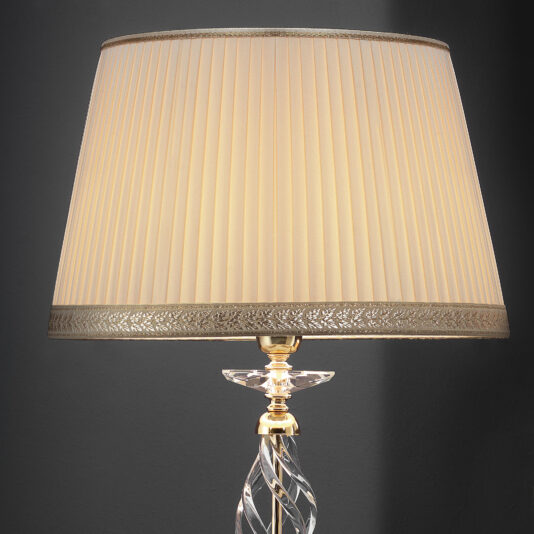 A close-up of the Twisted Table Lamp With Swarovski® Crystal, featuring a pleated beige lampshade adorned with intricate trim along the top and bottom edges. The lamp boasts an elegant twisted clear glass stem and a metallic base, set against a dark gray background.
