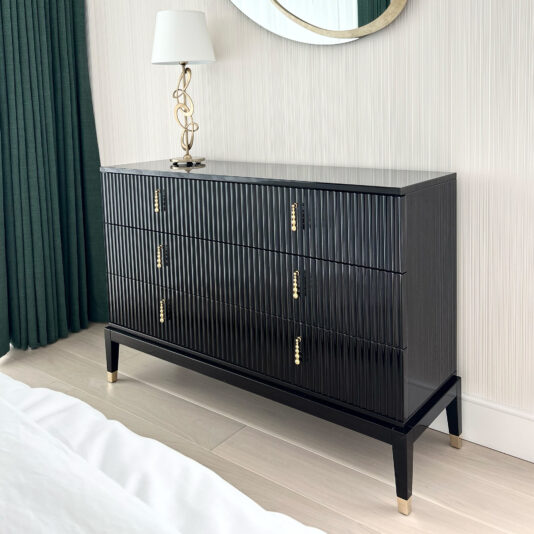 An Art Deco Style Chest of Drawers in black, featuring six vertical-grooved drawers with gold handles, stands against a light-colored wall. A lamp with a white shade and gold base sits on top of the chest. Behind the dresser is a green curtain, a round mirror with a gold frame, and light wooden flooring.