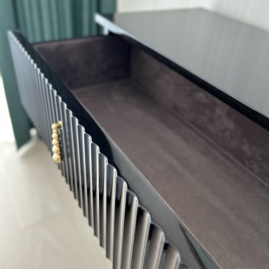 A close-up view of an open, empty drawer from the Art Deco Style Chest of Drawers reveals black, ribbed sides and a gold handle. The interior is lined with a dark, soft material. In the background, a light-colored floor and green curtain on the left hint at its elegant design.